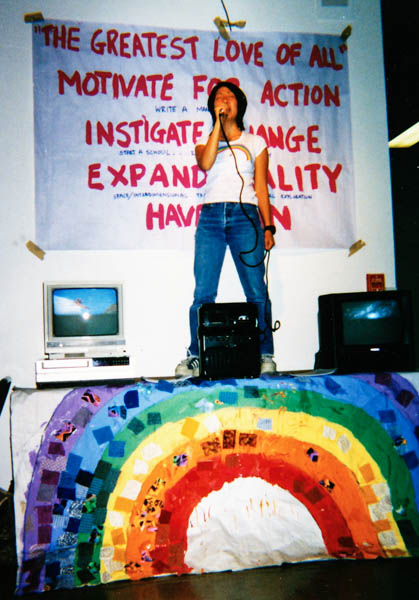 photo of Jennifer Moon standing on a table and singing into a microphone with a large poster containing text behind her and two TV monitors on each side, circa 1997
